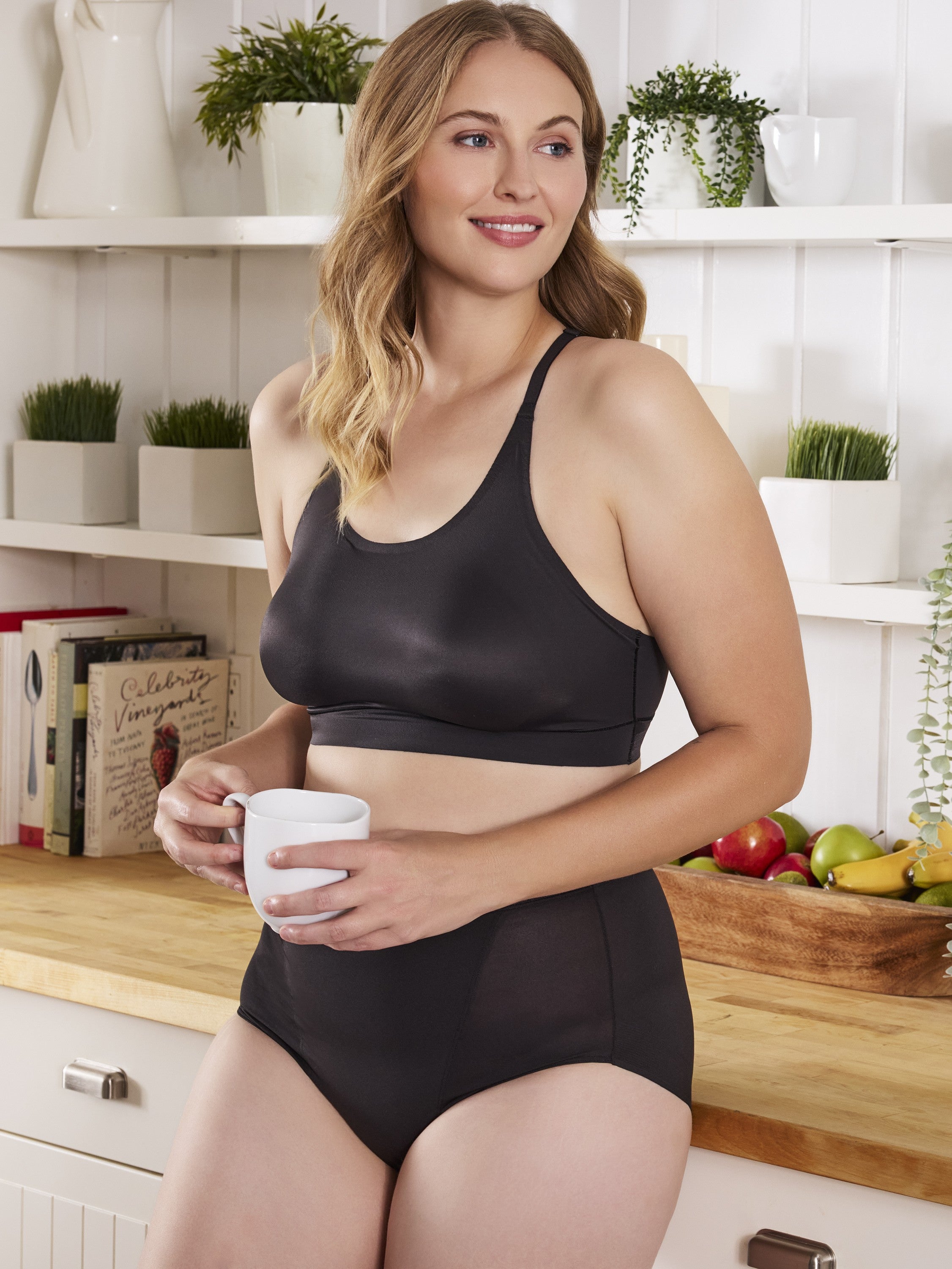 a woman standing in a kitchen holding a white coffee mug wearing a black bralette lounge bra and black control panties all-groups