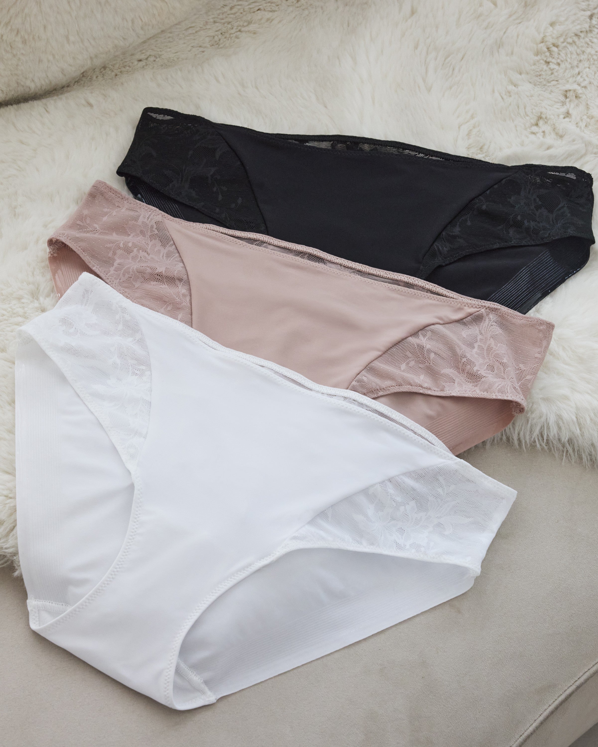 a black lace hipster panty, a rose pink lace hipster women's panty, and a white lace hipster women's underwear laying down on a fur blanket and suede sofa