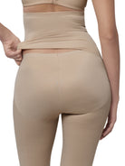 Rear view of a model wearing beige shapewear leggings and a matching high-waisted waistband top, lifting the waistband slightly with one hand against a white background.
