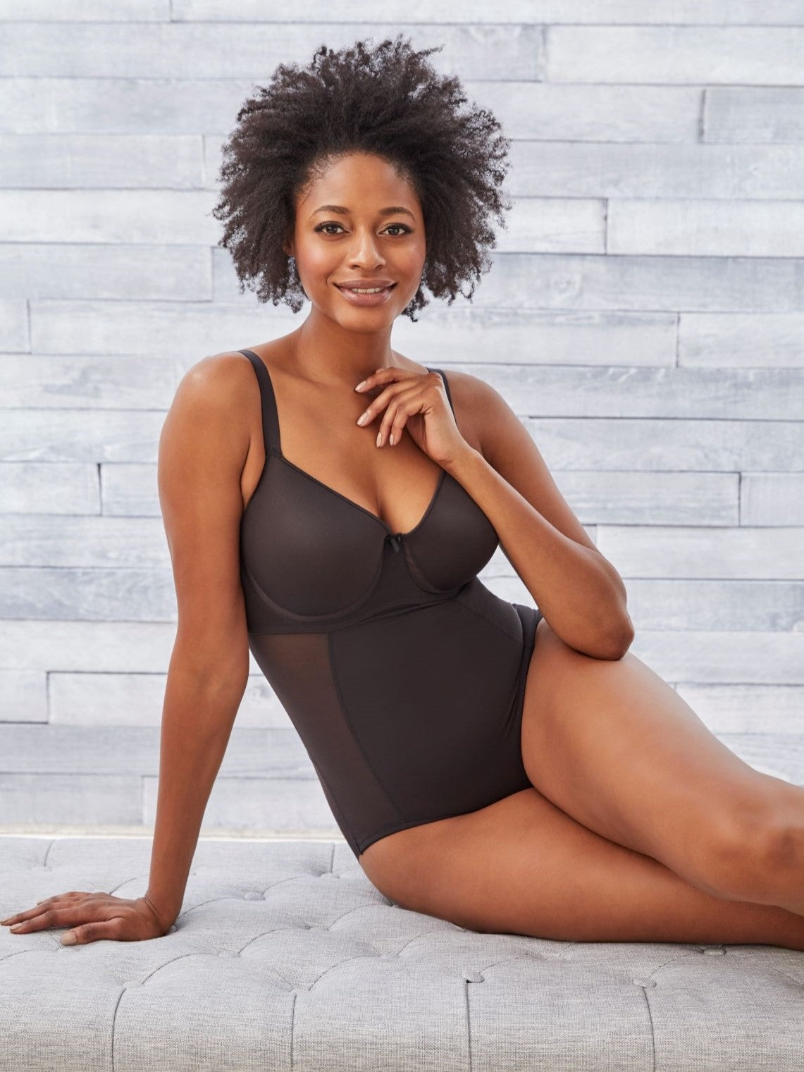 a woman sitting on a bench in front of a gray wall wearing a black tummy control body shaper shapewear bodysuit all-groups