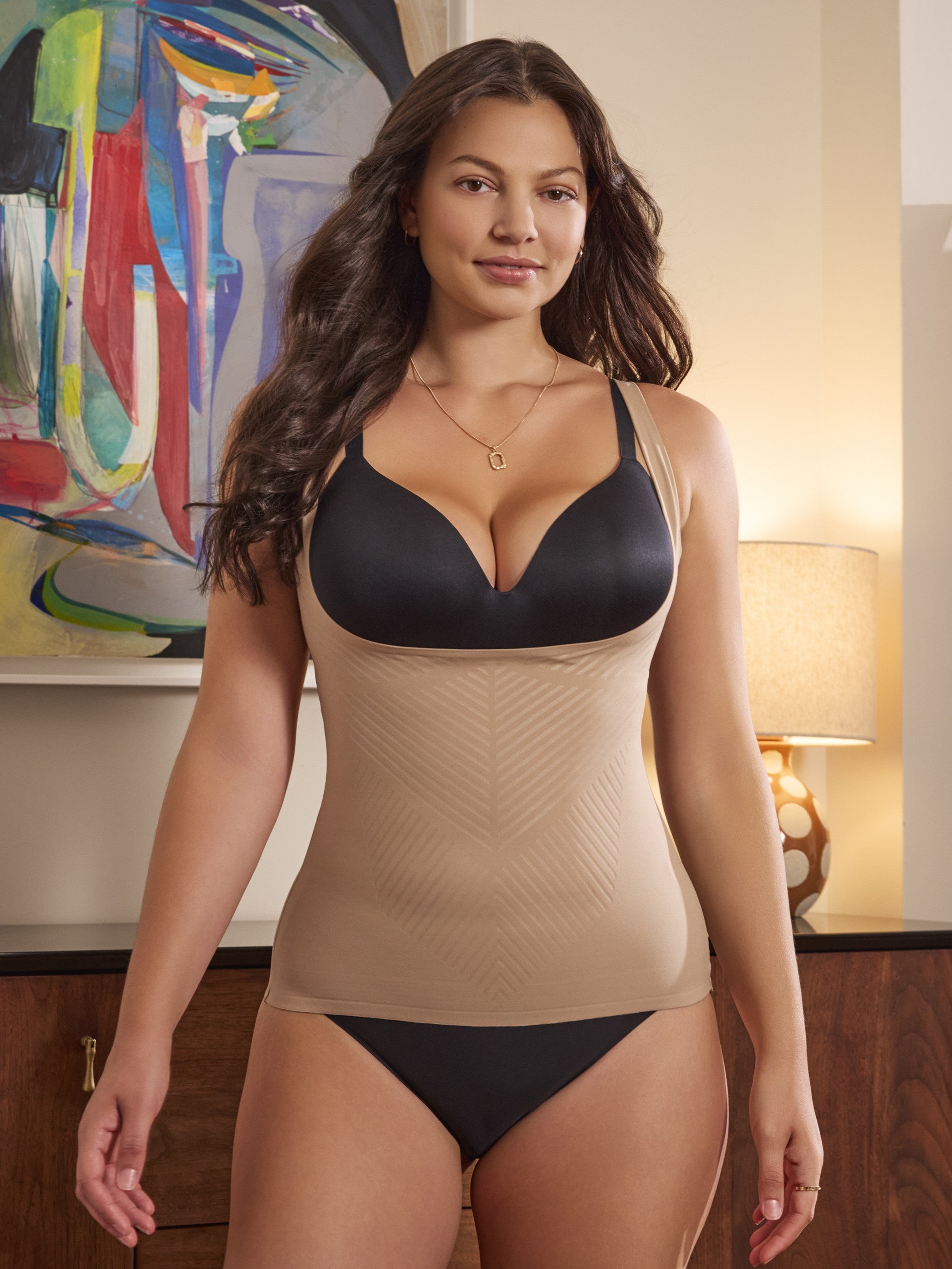 a woman standing in front of a colorful painting wearing a beige open bust shaping cami camisole shapewear tank top body shaper all-groups