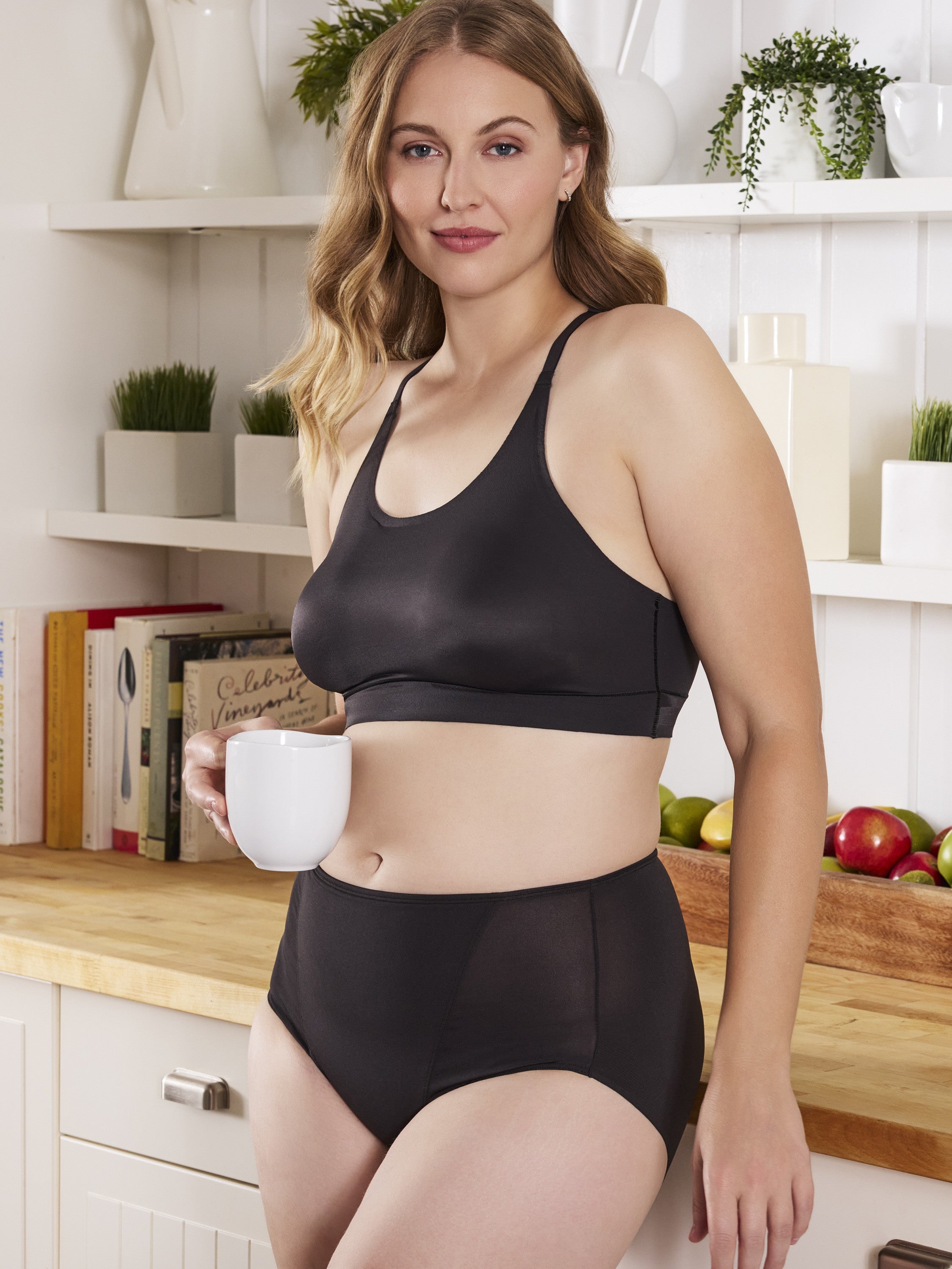 a woman leaning on a counter in a kitchen and holding a white coffee mug while wearing a black bra and a black tummy control top brief body shaper shapewear